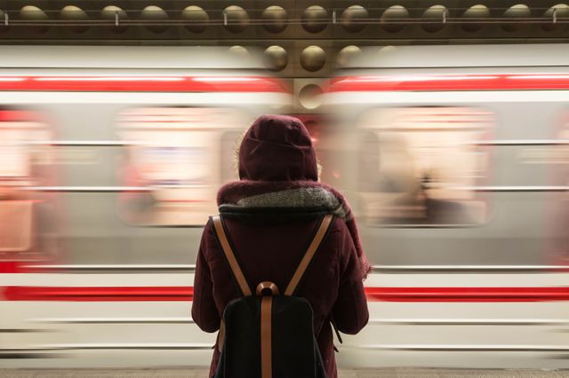 Person wearing a hooded jacket waiting on subway platform while train moves quickly. Useful for illustrating themes of urban life, daily commute, transportation, fast-paced environments, and travel.