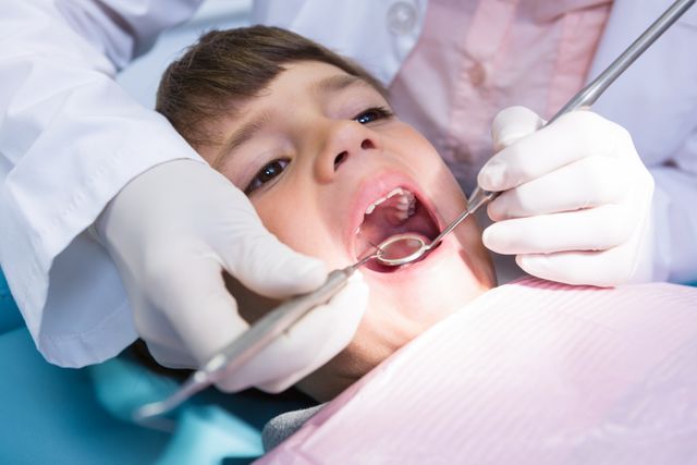 This image shows a dentist examining a young boy's mouth at a medical clinic. It is suitable for use in articles or advertisements related to pediatric dentistry, dental health, healthcare services, and child wellness. It can also be used in educational materials about the importance of regular dental checkups for children.