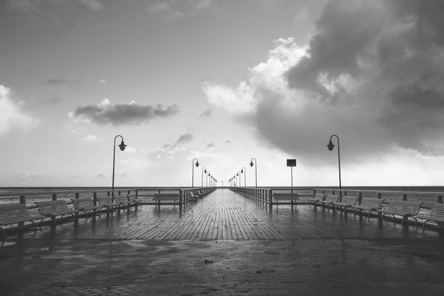 Empty wooden pier extends over calm ocean waters under an overcast sky. The symmetrical layout of benches and lampposts lining the boardwalk creates a sense of solitude and tranquility. Black and white tones enhance the moody atmosphere, making it perfect for use in themes related to calmness, reflection, isolation, seascapes, and travel inspiration. Ideal for website backgrounds, inspirational message visuals, and artistic prints.