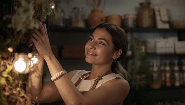 Young florist arranging hanging greenery and exposed bulbs in cozy boutique with warm glow
