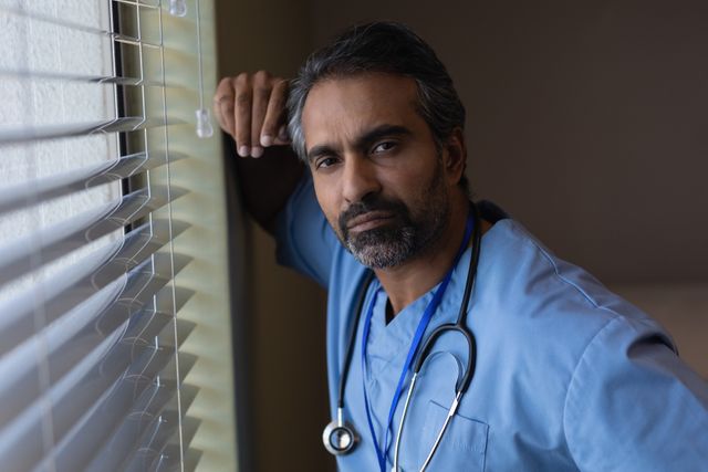 Portrait of biracial male surgeon standing near window at hospital