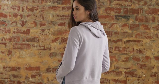 Young biracial woman stands against a brick wall, with copy space. Casual attire suggests a relaxed, urban setting.