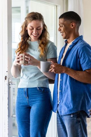 Smiling interracial couple standing in sunlit doorway holding coffee mugs morning chat