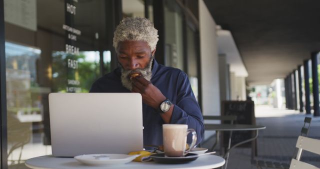 Senior man with grey beard working on laptop at outdoor café while eating breakfast and drinking coffee. Suitable for themes related to remote work, urban lifestyle, senior entrepreneurship, business, technology, or outdoor activities.