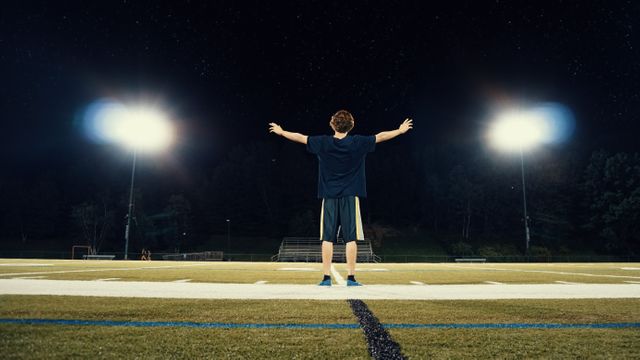 A man standing triumphantly with arms wide open on a sports field at night, under bright stadium lights. This can be used for concepts of success, sportsmanship, determination, and outdoor events.