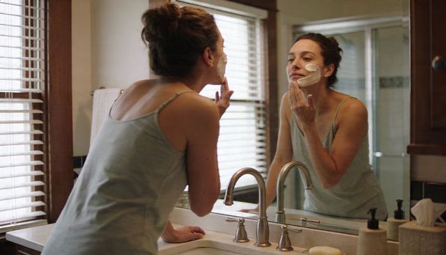 Woman applying moisturizer at bathroom vanity mirror during morning skincare routine