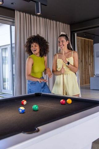 Multiethnic women laughing and playing pool in modern game room with black table and cues