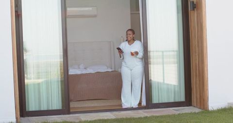 African american woman enjoying morning on balcony in pajamas holding smartphone and mug