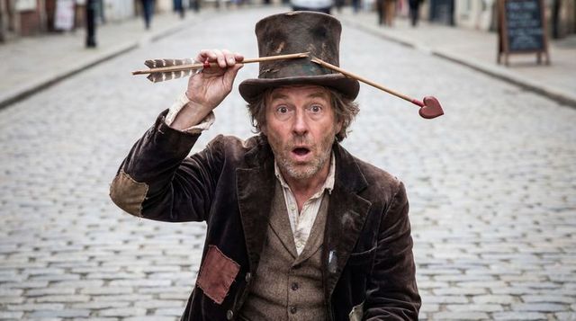 Street performer balancing heart-tipped arrow on top hat wearing patched vintage jacket
