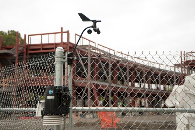 This image shows an air sampler mounted on a fence at a construction site. The structure in the background is the NASA Ames Green Building, also known as Sustainability Base, located at Moffett Field, CA. This picture depicts the emphasis on environmental monitoring and sustainable building practices. Suitable for use in articles about construction technology, sustainable architecture, eco-friendly building projects, and research centers.