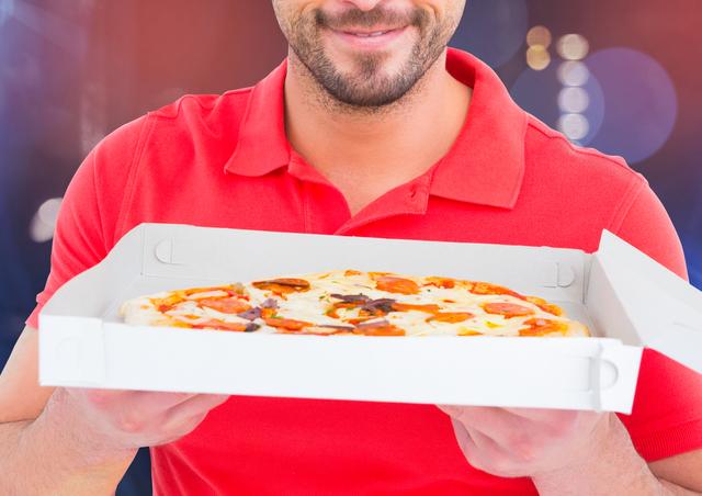 Smiling delivery man holding a freshly baked pizza in an open box. He is wearing a red polo shirt, which typically represents food delivery uniforms. The background has a bokeh effect with colorful lights, indicating an urban setting during evening time. Useful for illustrating food delivery services, online ordering, fast food promotions, and customer satisfaction in the food industry.