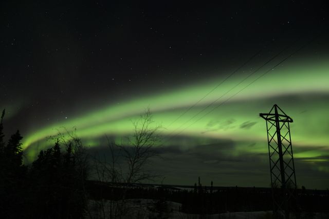 Beautiful view of northern lights illuminating the dark sky over a snowy forest. Electric tower in foreground emphasizes vast wilderness. Suitable for use in nature-themed projects, travel brochures, wallpapers, and educational materials about auroral phenomena.
