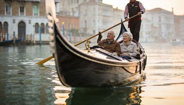 Senior couple enjoying venice sunset gondola ride, smiling gondolier steering ornate boat