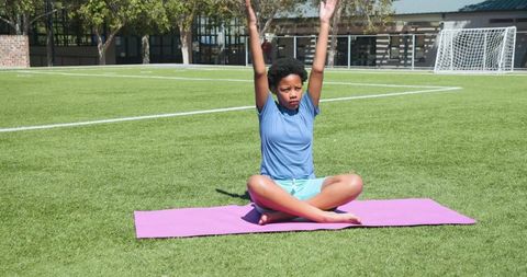African adolescent girl practicing yoga cross-legged on soccer field on pink mat