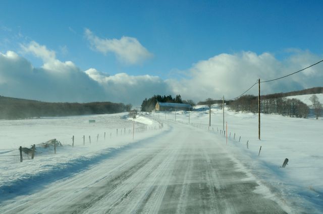 Snow-covered road extending through a winter countryside on a clear day with blue sky and fluffy clouds. Ideal for depicting seasonal changes, winter travel scenes, country life, serene nature backgrounds, or rural beauty.