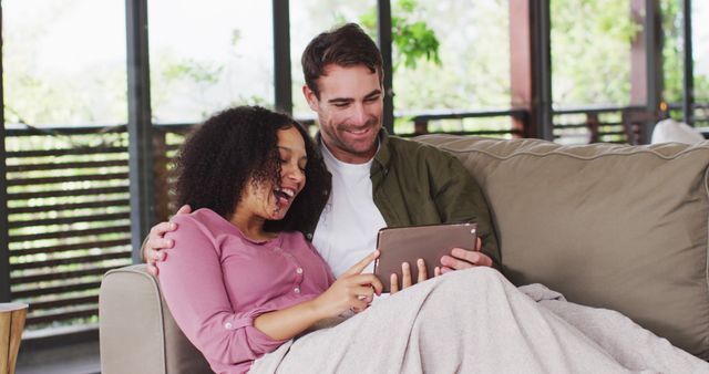 Couple sitting closely on sofa, both are enjoying and smiling while looking at tablet screen. Bright indoor setting with natural light coming through large windows. Ideal for use in lifestyle blogs, adverts focused on home entertainment, smartphone or tablet promotions, or articles about technology affecting family life.