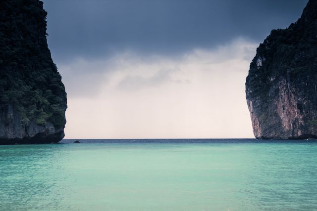The image captures a tranquil tropical beach framed by two majestic limestone cliffs with calm turquoise water extending to the horizon under a slightly overcast sky. Ideal for travel advertisements, tourism brochures, and websites promoting vacation destinations. It effectively evokes feelings of relaxation, natural beauty, and the exotic allure of tropical getaways.