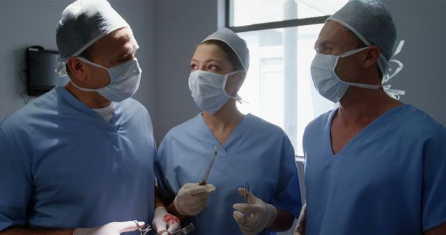 Three surgeons in blue scrubs are standing in an operating room, engaged in conversation. They are wearing surgical masks and gloves, and holding medical instruments, indicating a discussion about a surgical procedure. This image can be used to illustrate team collaboration, medical procedures, and healthcare environments in educational materials, medical articles, or hospital brochures.