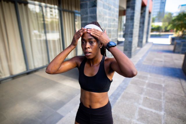 This image shows a tired African American woman walking outdoors after a workout. She is wearing sportswear and appears to be recovering from an intense exercise session. This image can be used for promoting fitness, healthy lifestyles, workout routines, and urban exercise environments.