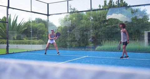 Female teammates playing padel on blue court with glass walls and lush green backdrop