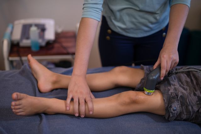 Therapist using ultrasound machine on child's knee in hospital ward. Useful for illustrating medical treatments, pediatric care, physical therapy, and healthcare services.