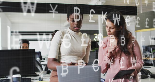 Two professional women discussing a project at a modern office desk with digital financial symbols overlay (currency signs and technical graphics), emphasizing the intersection between business management and financial technology. Suitable for themes on finance, teamwork, diverse workplace, digital transformation, fintech, or collaboration.