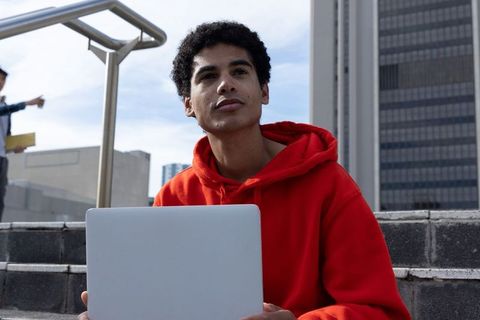 African american teen working on laptop outdoors on urban steps wearing red hoodie