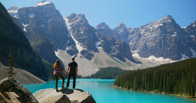 Caucasian tourist couple standing on cliff, holding hands and looking at mountains by sunny lake. Summer, vacations, traveling and active lifestyle, unaltered.