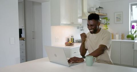 African american man celebrating work win while using laptop at bright modern kitchen island