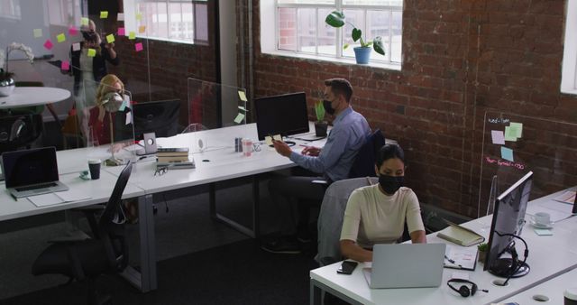 Diverse group of employees working in a modern office environment, wearing masks for safety. Contemporary office features brick walls, large windows, and open space with workstations equipped with computers and office supplies. Photo is ideal for illustrating workplace safety, pandemic measures, modern office culture, and teamwork.