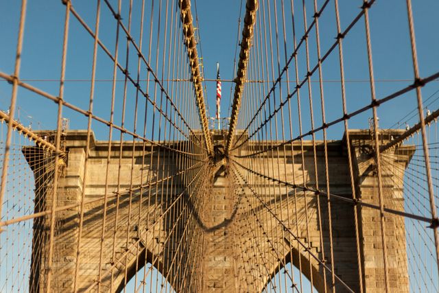 Capturing cables and tower of Brooklyn Bridge from underneath against clear blue sky, this image highlights the intricate design and architectural details of the famous bridge. Ideal for travel blogs, architecture and engineering articles, New York City tourism promotions, and educational materials about historic landmarks. The absence of people emphasizes the grandeur and scale of the structure.
