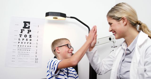 The boy is joyfully high-fiving the optometrist after a successful eye exam in a clinic. The boy is smiling and wearing glasses, indicating satisfaction with the exam result. The background includes an eye chart. This imagery is ideal for advertising pediatric healthcare clinics, optometry services, and promoting positive patient experiences in medical settings.