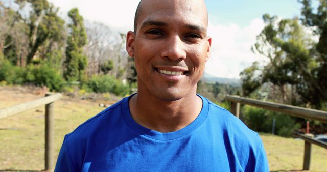 A young African American man smiles at the camera, wearing a blue shirt and standing outdoors with trees and a fence in the background, with copy space. His cheerful expression and the sunny environment contribute to a positive and uplifting atmosphere.