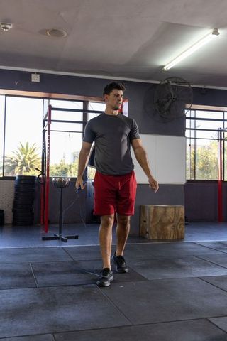 Fit man walking with jump rope in gym wearing gray shirt and red shorts