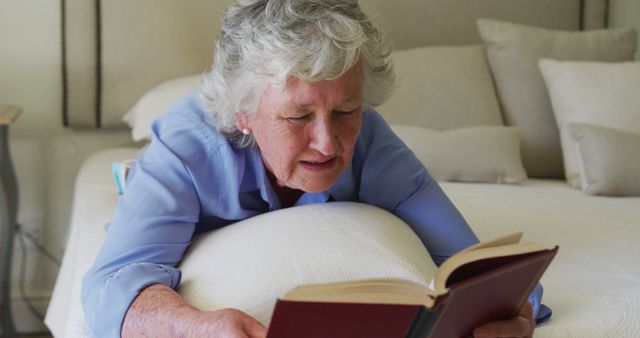 Elderly woman with grey hair wearing blue shirt is lying on a cozy bed while deeply engrossed in a book. Ideal for themes involving leisure, relaxation, senior lifestyle, home comfort, quiet moments, mental engagement, or peaceful environment. Can be used for lifestyle blogs, elder care services, retirement living promotions, or public health campaigns focused on mental wellness and reading for seniors.