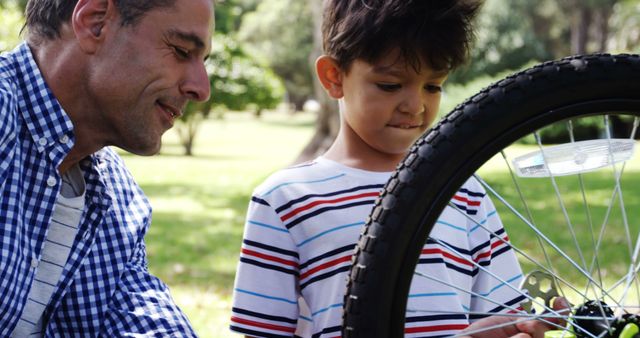 Father shows son how to repair bicycle in park, fostering learning and bonding. Suitable for themes of family activities, parenting, outdoor fun, hands-on learning, and father-son relationships.