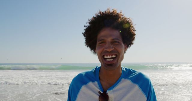African American man standing on a beach, smiling with ocean waves in the background. Suitable for travel brochures, summer promotions, beach vacation advertisements, or lifestyle blogs focusing on leisure and relaxation.