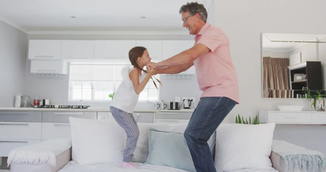 Happy caucasian grandfather and granddaughter holding hands and jumping on sofa. Lifestyle, domestic life, family, and togetherness.
