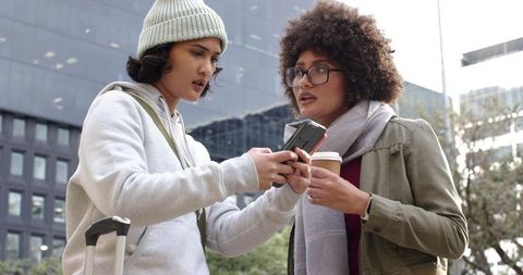 Young women sharing directions on smartphone in city plaza holding luggage and coffee cup