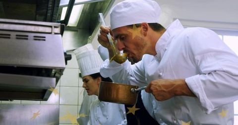Professional chef tasting sauce with ladle and copper saucepan in busy restaurant kitchen