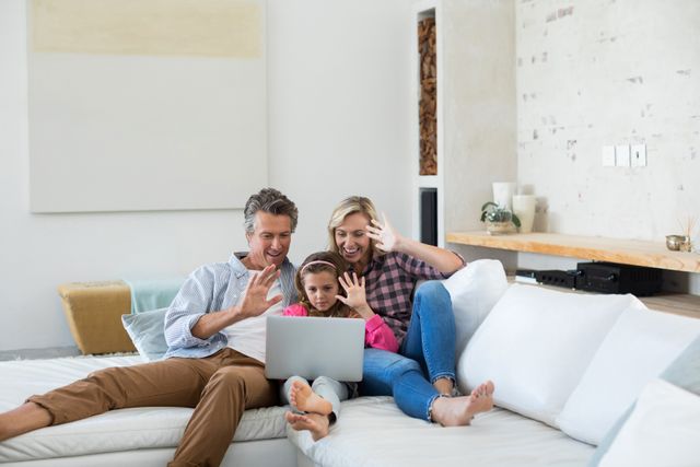 Family sitting on a comfortable couch in a modern living room, engaging in a video call on a laptop. They are smiling and waving, showcasing togetherness and the use of technology for communication. Ideal for use in advertisements, articles, or blogs about family bonding, modern technology, remote communication, and home life.