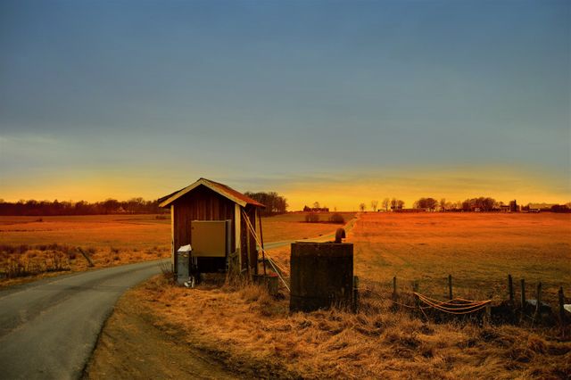 A serene rural sunset over open fields with a small wooden shed, showcasing the tranquil countryside atmosphere. This image captures the simplicity and beauty of farm life with golden light casting hues over the landscape. Perfect for use in projects related to agriculture, rural living, tourism, and inspirational nature content. The quiet and peaceful mood is ideal for backgrounds, promotional materials, and websites focusing on outdoor activities and eco-friendly living.