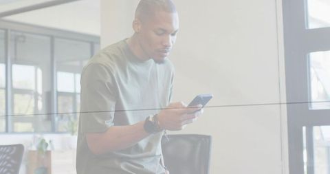 Young man leaning on glass partition checking smartphone in modern office workspace