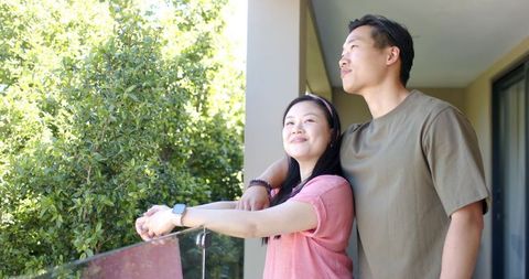 Asian couple enjoying sunlit balcony, leaning on glass railing, woman wearing smartwatch