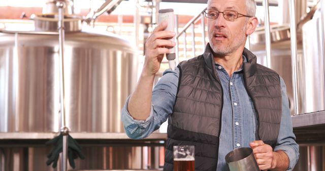 This stock photo depicts a senior male brewer inspecting the quality of beer in a brewery. He is holding a device to check certain qualities of the beer, showcasing professionalism and attention to detail in the brewery environment. This image is ideal for use in articles or advertisements related to craft beer, brewery tours, or brewing education. It highlights the expertise and dedication required in the brewing industry.