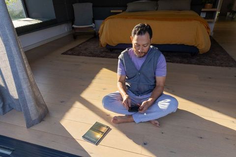 Mature asian man relaxing on bedroom floor using smartphone in sunlit minimalist home