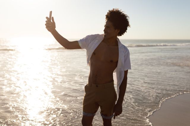 Young man enjoying a sunny day at the beach, taking a selfie with his mobile phone. Ideal for use in travel promotions, summer vacation advertisements, lifestyle blogs, and social media content.