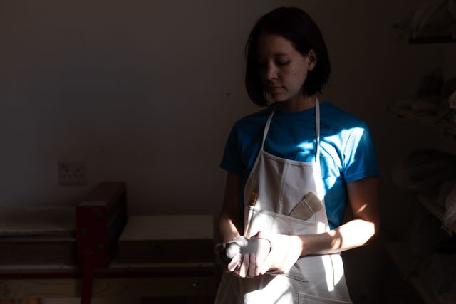 Front view of a Caucasian female potter with dark red hair in a bob hairstyle, holding in hands a lump of clay in a pottery studio, with equipment in the background, wearing white apron.