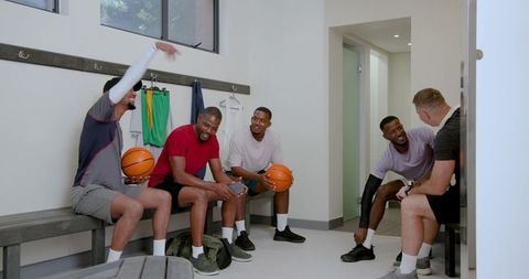 Diverse male teammates chatting in locker room with basketballs showing camaraderie