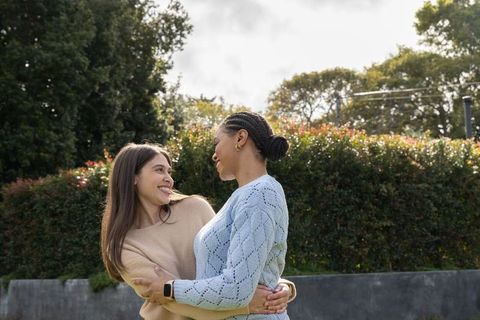 Two women embracing and smiling in garden wearing cozy sweaters, gold hoops, smartwatch